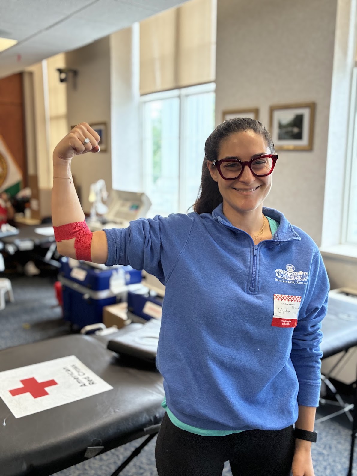 A person in a blue sweatshirt with a name tag flexes her arm, which is wrapped with red bandage indicating a recent blood donation. Behind her is a soft table with the American Red Cross logo.