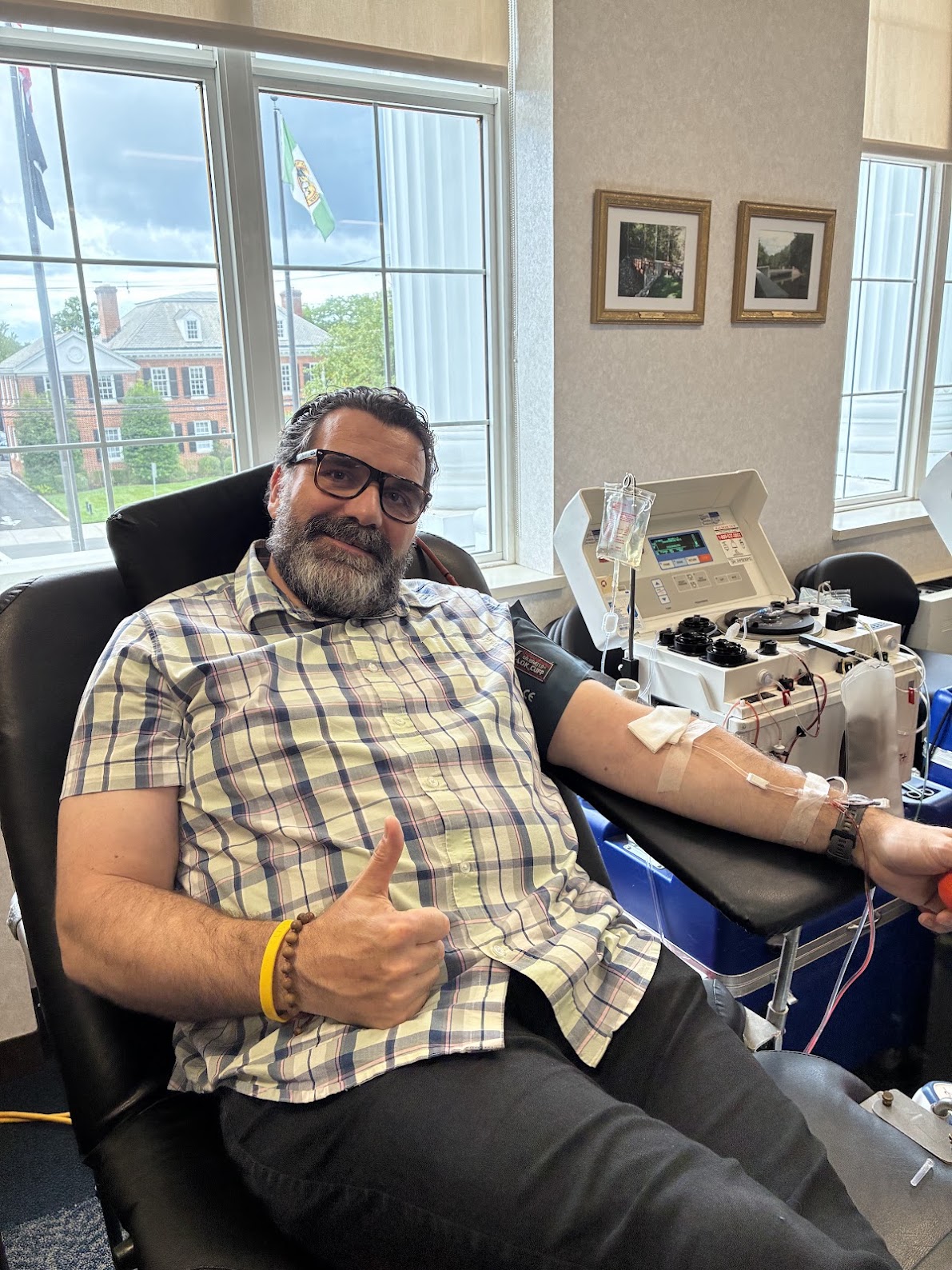 A person sites on a table with a tube connected to his arm and a machine behind him. He sticks his thumb up as he waits for his blood donation to finish.