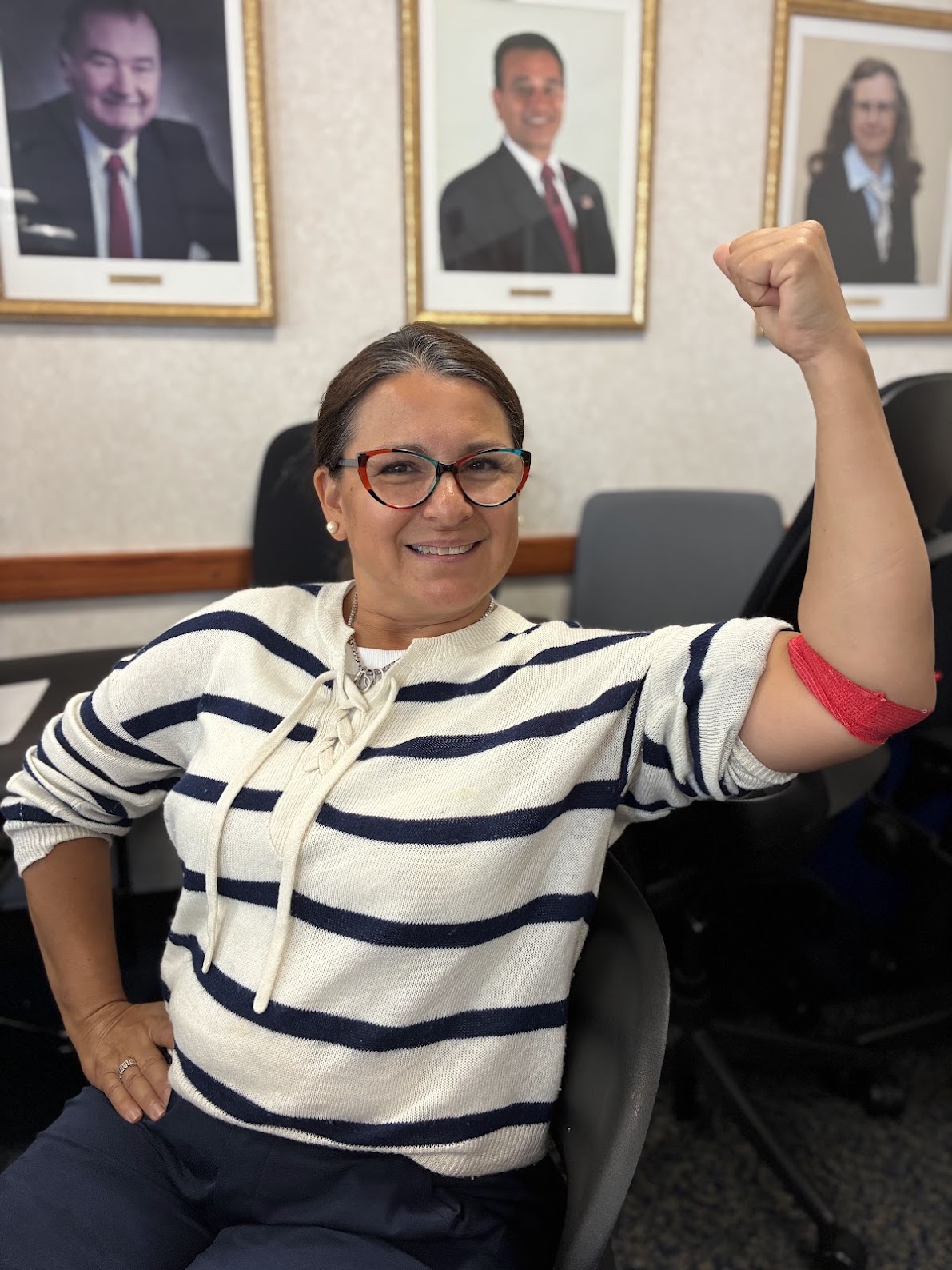 A person in a blue and white striped sweatshirt flexes her arm which is wrapped in red bandage indicating a recent blood donation. There are portraits of people behind her.