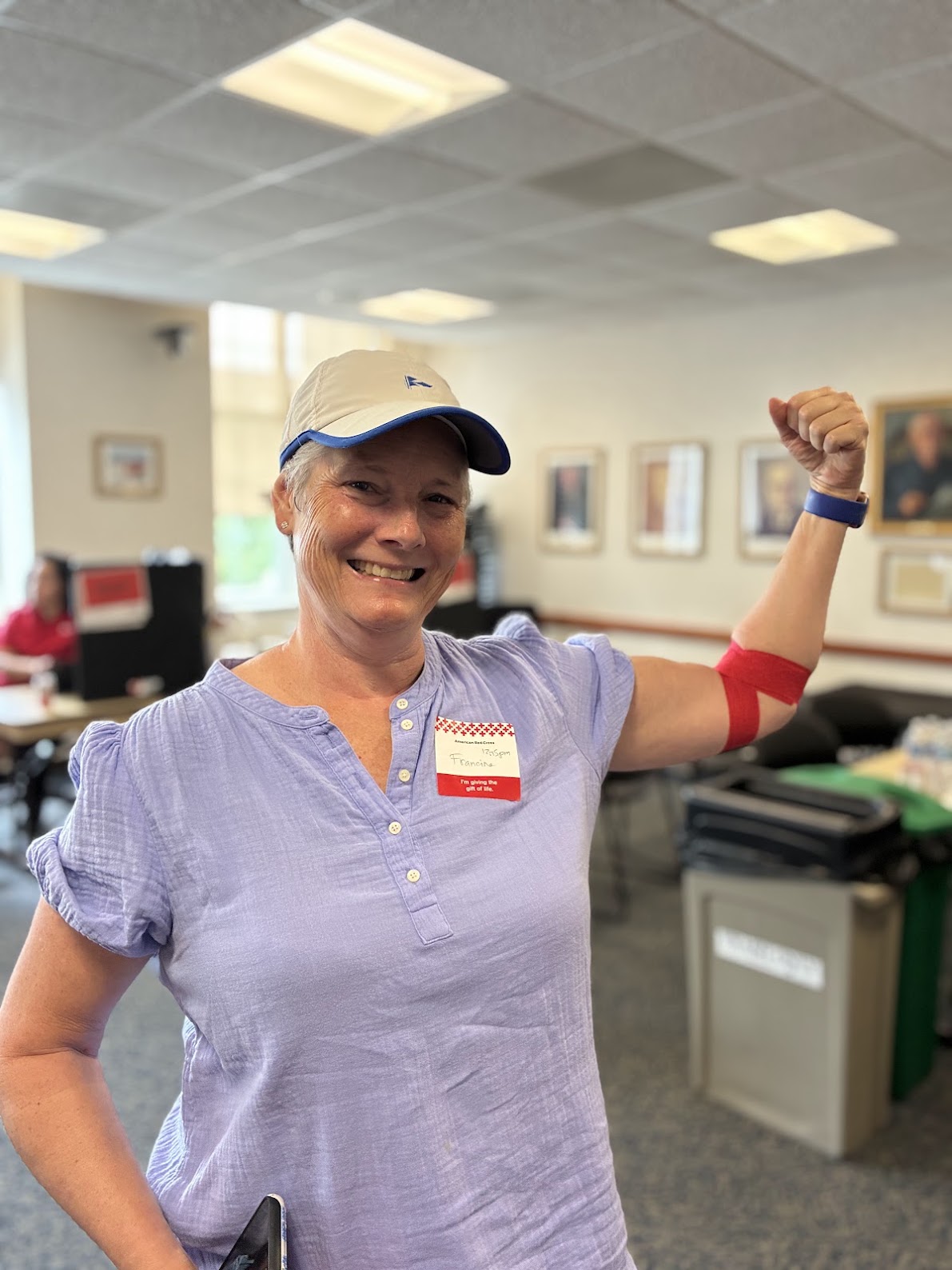 A person in a purple shirt with a name tag flexes her arm, which is wrapped with red bandage indicating a recent blood donation. 