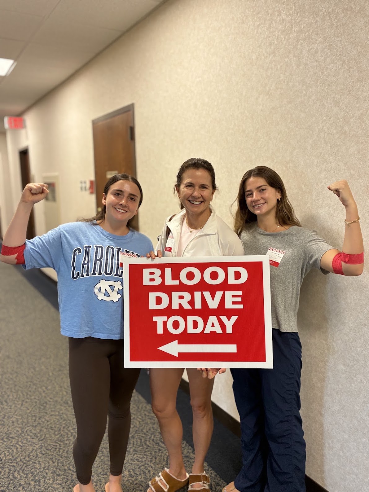 Three people stand together in a hall, holding a sign that says Blood Drive Today, with the outer two younger people flexing their arms that are wrapped with red bandage indicating a recent blood donation.