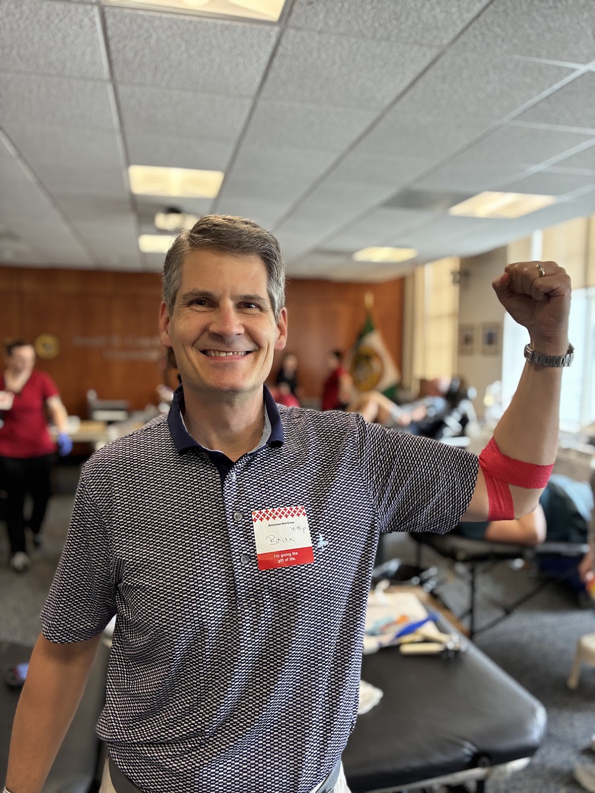 A person in a blue patterned shirt with a name tag flexes his arm, which is wrapped with red bandage indicating a recent blood donation. 