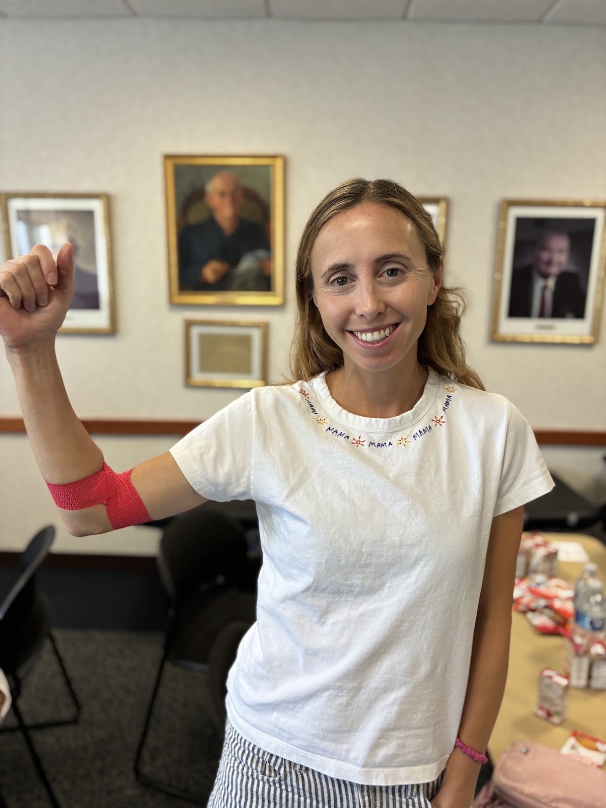 A person in a white shirt that has “mama” written repeatedly around the collar flexes her arm, which is wrapped with red bandage indicating a recent blood donation. 