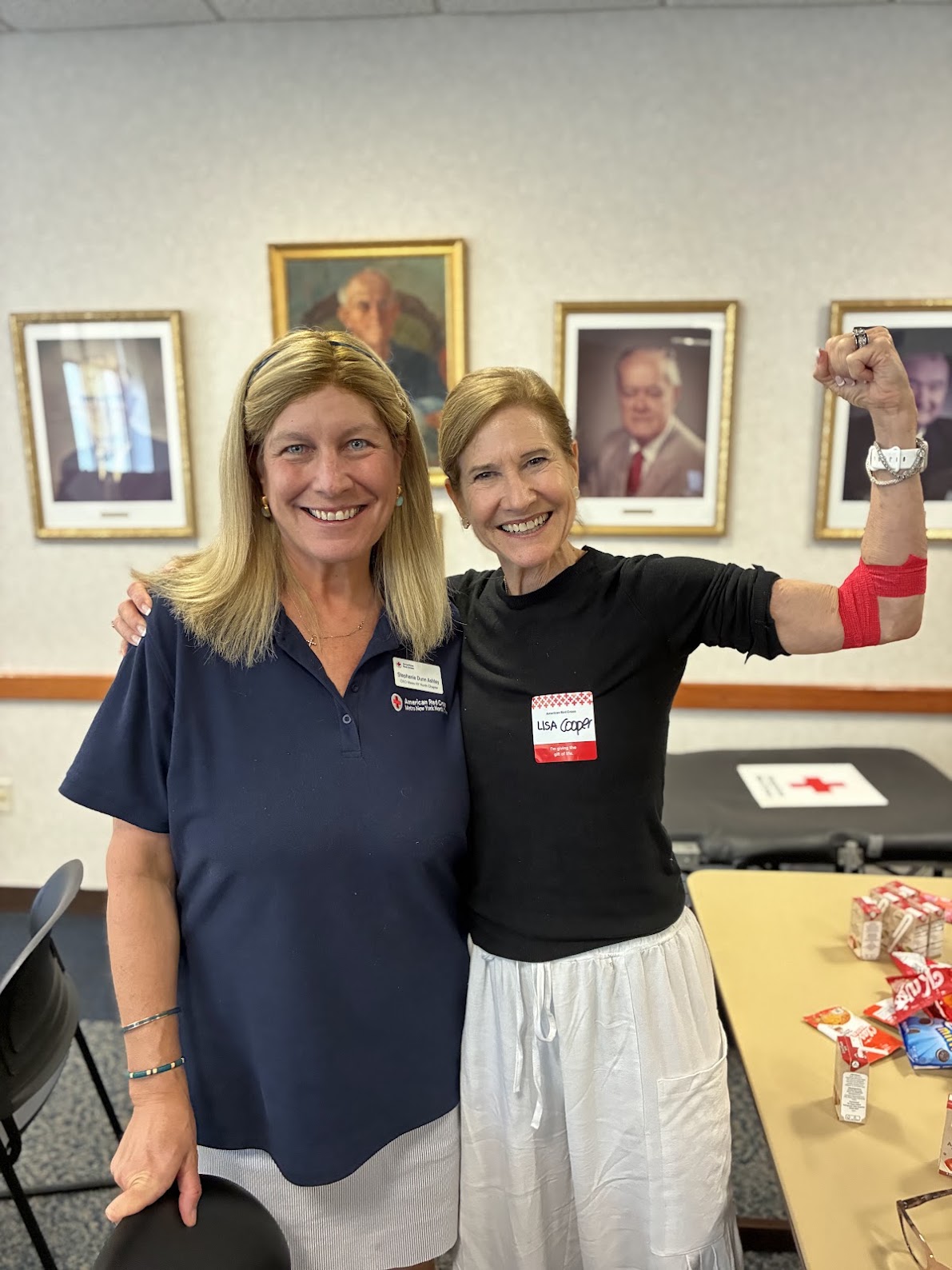 Two people stand together, with one person wearing a name tag and flexing an arm wrapped with red bandage indicating a recent blood donation. There is a soft black table behind them with the Red Cross logo.