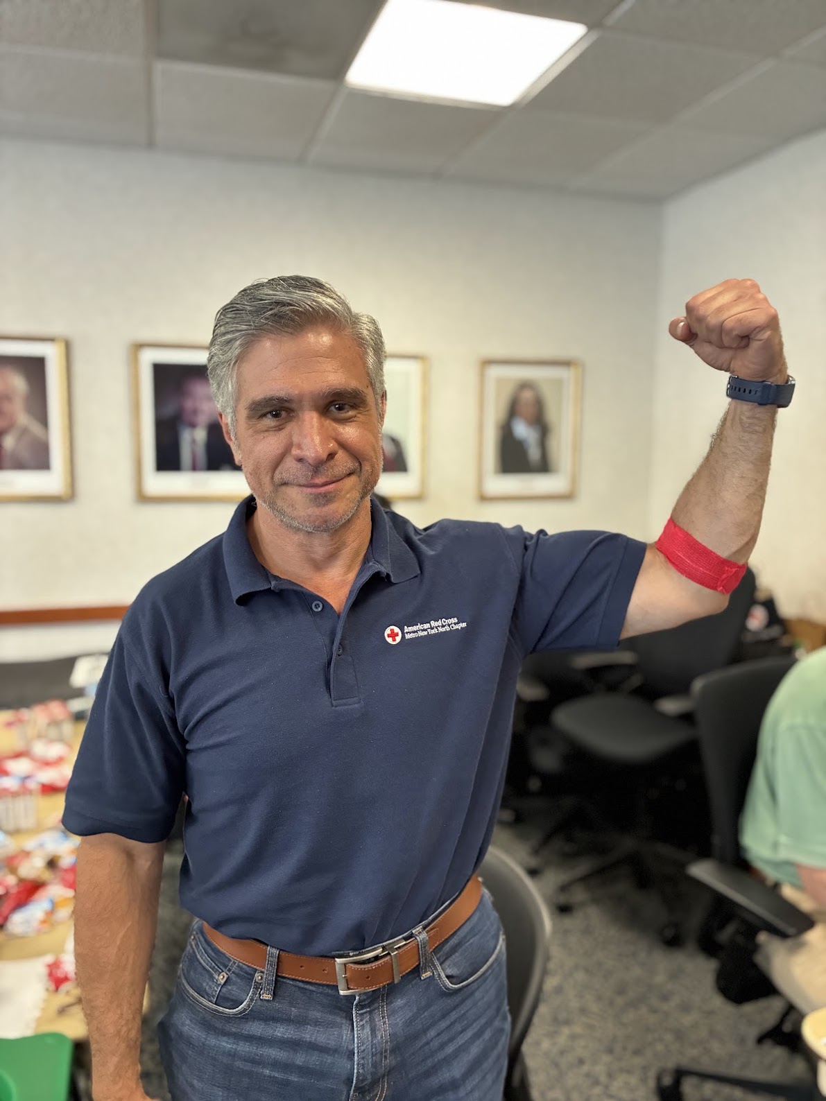 A person in a blue shirt with a Red Cross logo flexes his arm, which is wrapped with red bandage indicating a recent blood donation. 