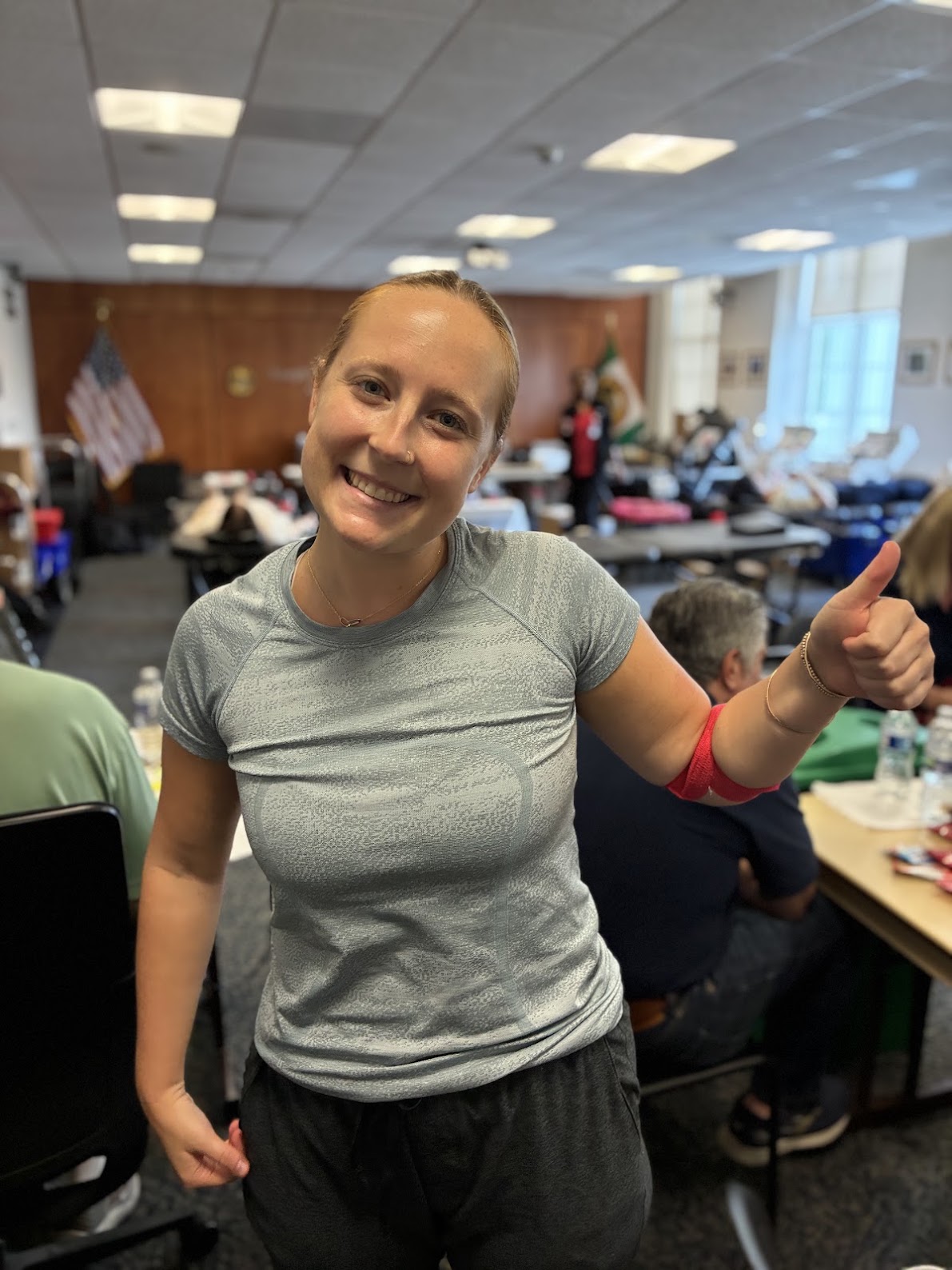 A person in a grey shirt flexes her arm, which is wrapped with red bandage indicating a recent blood donation. Behind her are a series of tables set up for blood donations.