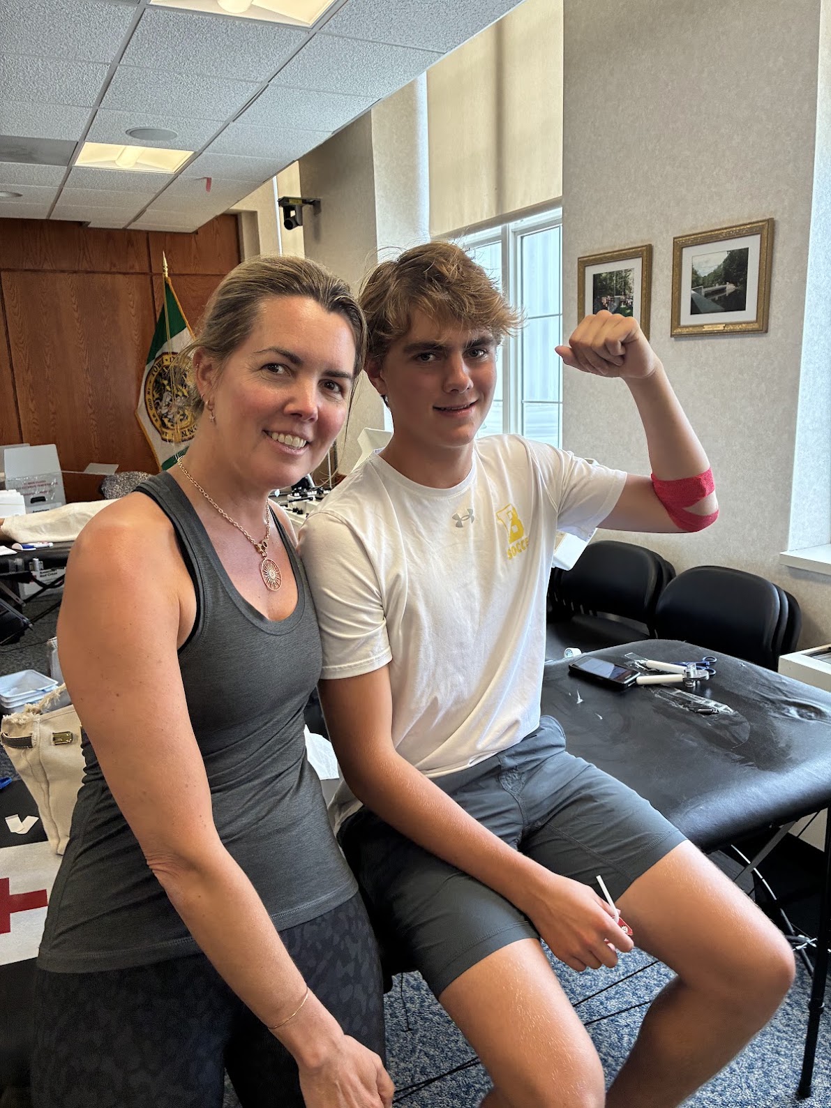 Two people sit on a table together, the younger person on the right flexes his arm which is wrapped in a red bandage indicating a recent blood donation.
