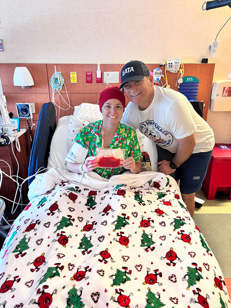 Hope Hall in a hospital bed holding a bag of donated blood with her husband, Bo Hall standing beside her.
