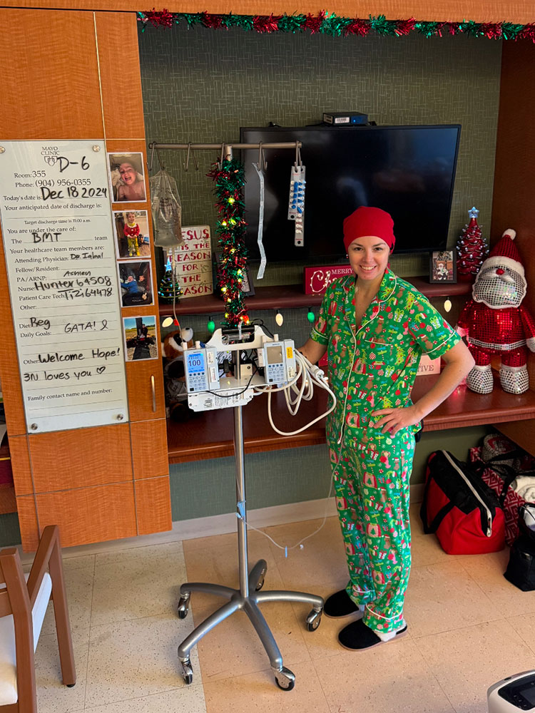 Hope Hall in the hospital smiling, wearing pajamas, a head wrap and standing next to medical equipment.