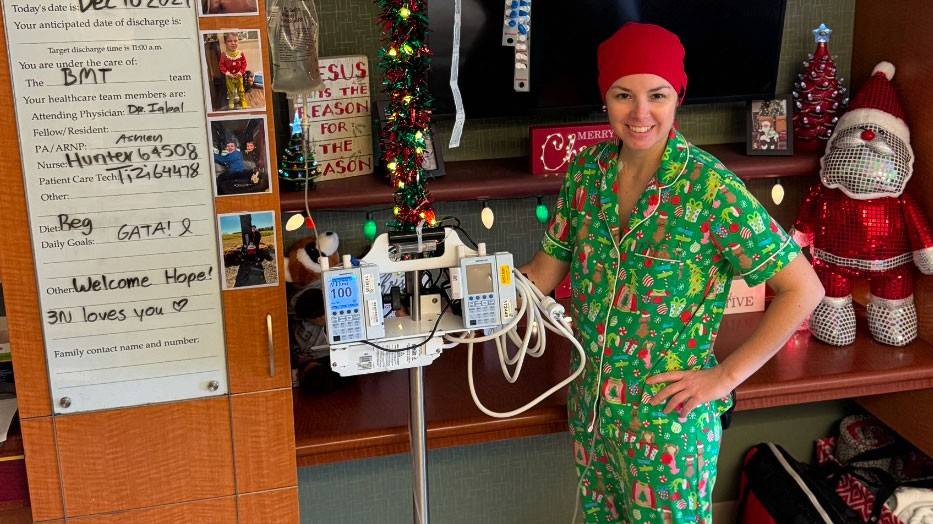 Hope Hall in the hospital smiling, wearing pajamas, a head wrap and standing next to medical equipment.