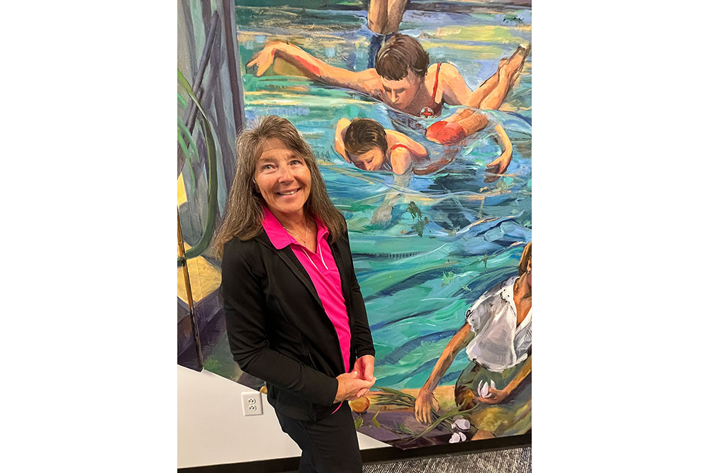 Birch Noden standing next to a mural of Red Cross swim instructor training a child.