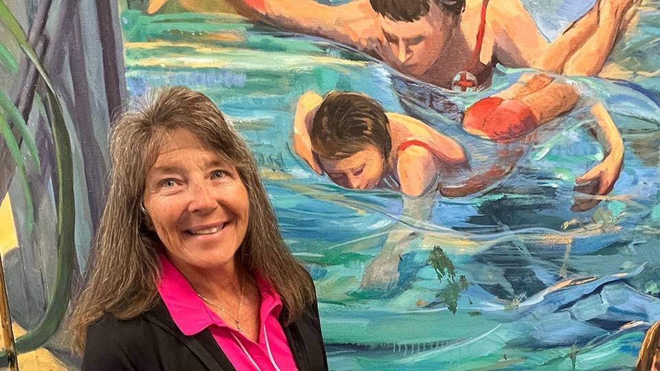 Birch Noden standing next to a mural of Red Cross swim instructor training a child.