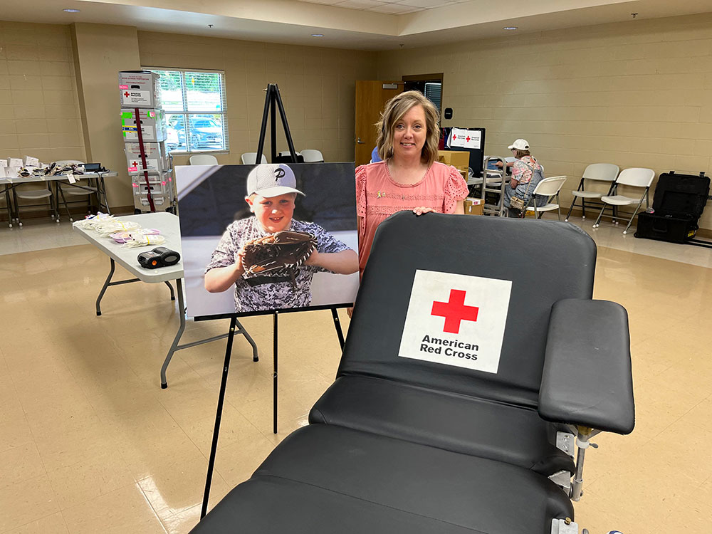 Kimberly Millsap stands beside an American Red Cross donation chair, smiling. An easel displays a photo of Blake Millsap with a baseball glove.