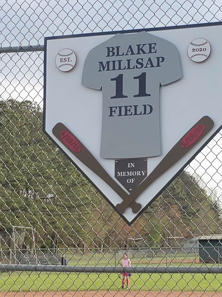 In memory of sign for "Blake Millsap Field" with a jersey, two bats, and dates. A child in pink plays on the field behind a chain-link fence.