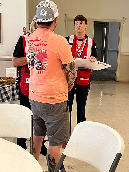 a Red Cross volunteer holding a clipboard and speaking to a man in a shelter.