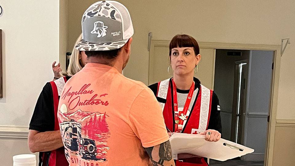 a Red Cross volunteer holding a clipboard and speaking to a man in a shelter.