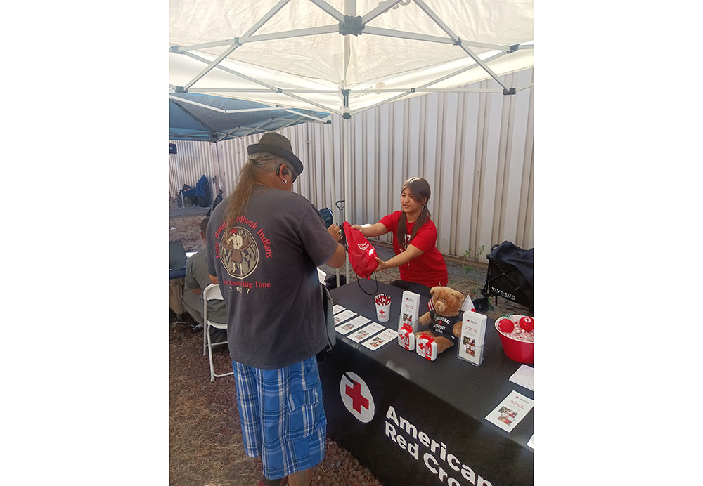 A man receives a red bag from a smiling volunteer, Anna Maxwell, at an American Red Cross booth. The table displays pamphlets, pens, and a stuffed toy under a canopy.