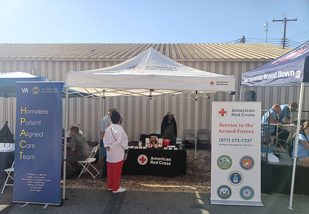 Outdoor event with American Red Cross and VA booths. People interact under canopies promoting services for veterans and the homeless. Sunny day, community-focused.
