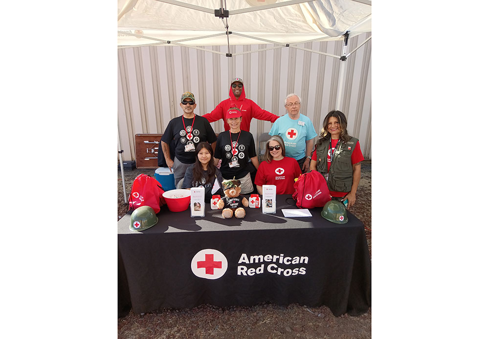 A group of seven volunteers stand and sit under a canopy at an American Red Cross booth. They are smiling, wearing shirts with Red Cross logos, and surrounded by branded items and a teddy bear on the table, creating a welcoming and supportive atmosphere.