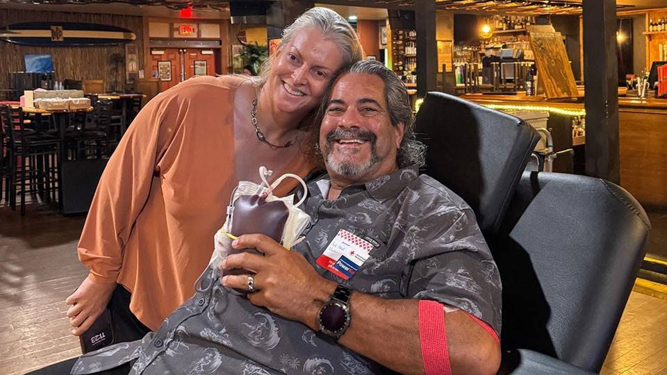 Derek Clark sitting in a blood donation chair holding a bag of donated blood while his wife leans over next to him.