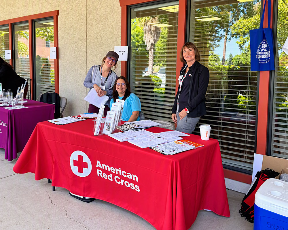 three Red Cross volunteers behind a table with Red Cross promotional items on it.