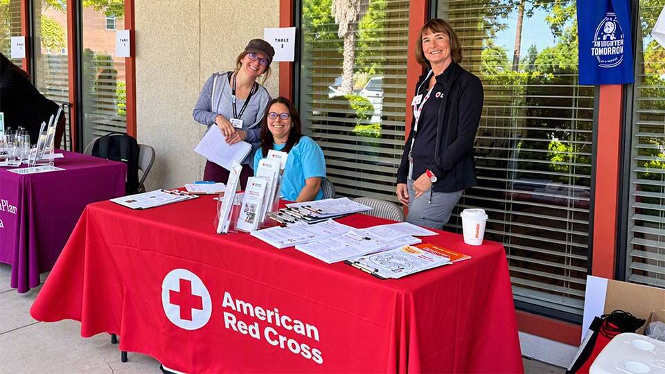 three Red Cross volunteers behind a table with Red Cross promotional items on it.