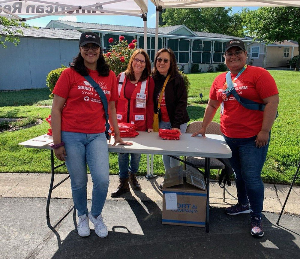 four Red Cross volunteers standing at a table in a parking lot with Red Cross promotional items.