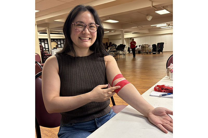 a woman, wearing glasses, sitting in a chair and pointing at her red bandage around her arm.