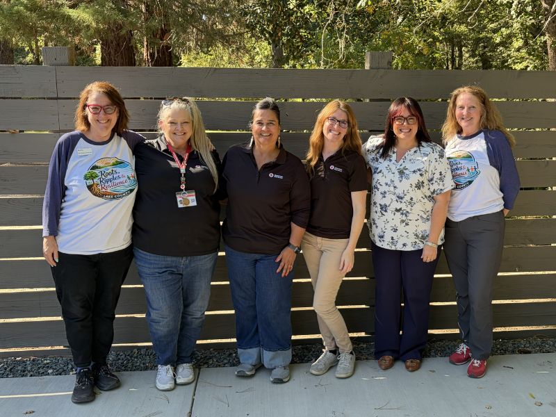 6 smiling women stand in front of fenced area with trees