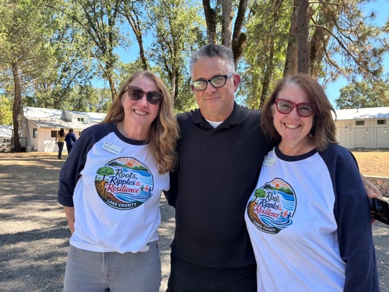 three people smiling wearing a "roots, ripples, and resilience" t-shirt