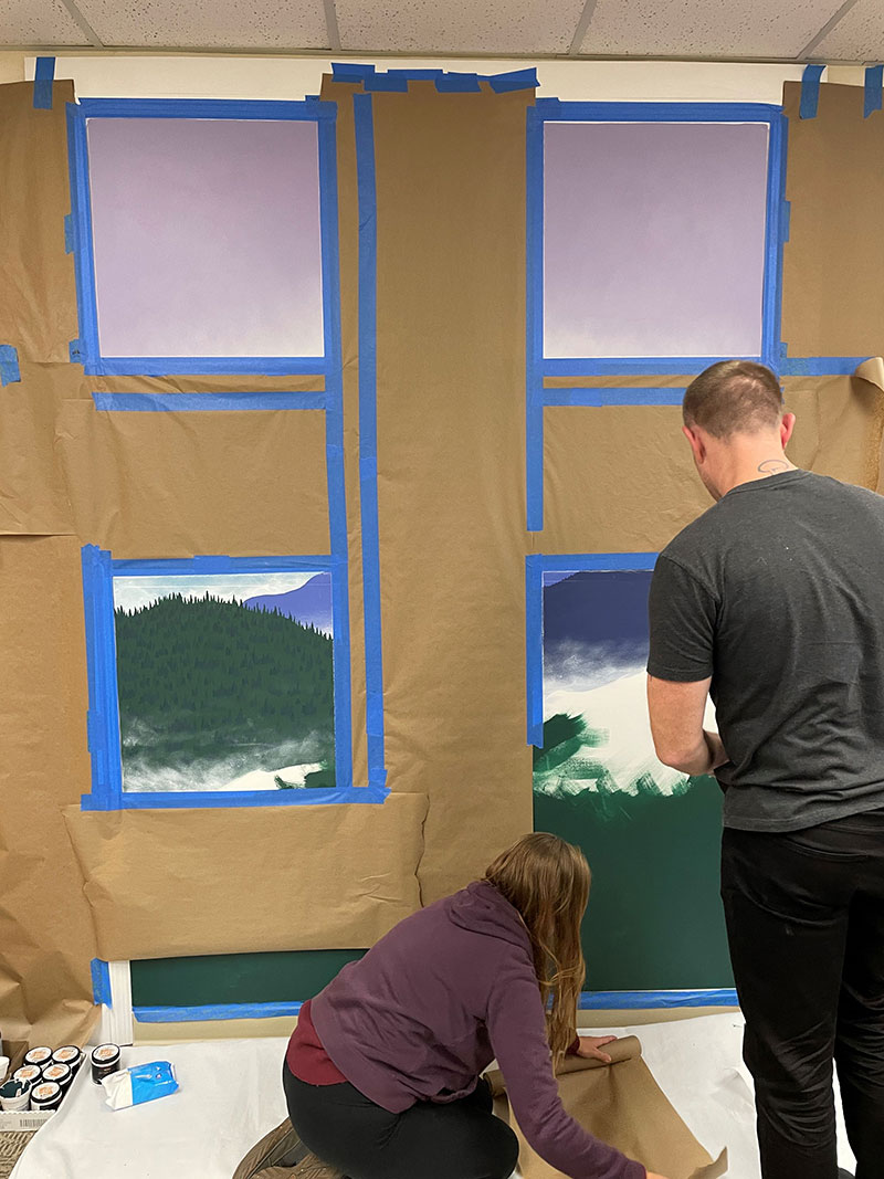 Two people painting a mural that is partially covered in brown paper and masking tape.