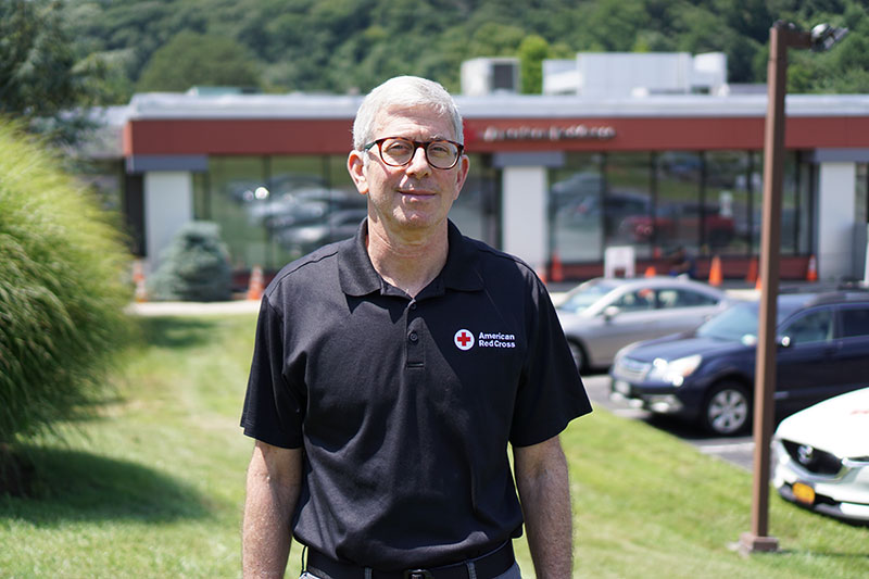 Alec Cecil wearing glasses and a Red Cross shirt while standing on grass next to a parking lot on a sunny day.