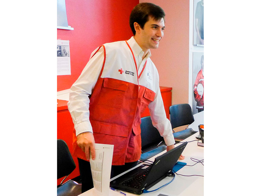 Red Cross volunteer Brandon Sherr wearing a Red Cross vest and standing in front of a desk with a laptop computer.