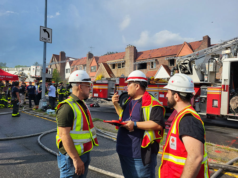 Three American Red Cross volunteers in vests and hard hats with fire fighters, fire engine and houses in the background.