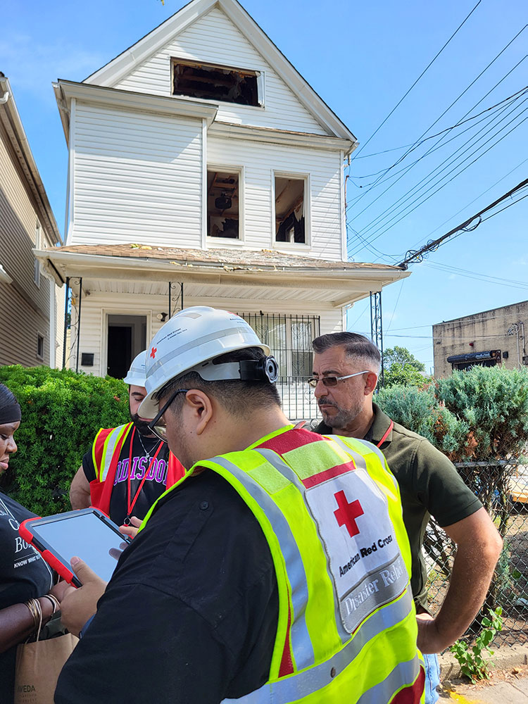 Two American Red Cross volunteers in vests and hard hats talking with two people and a two story house in the background that has been damaged by fire.