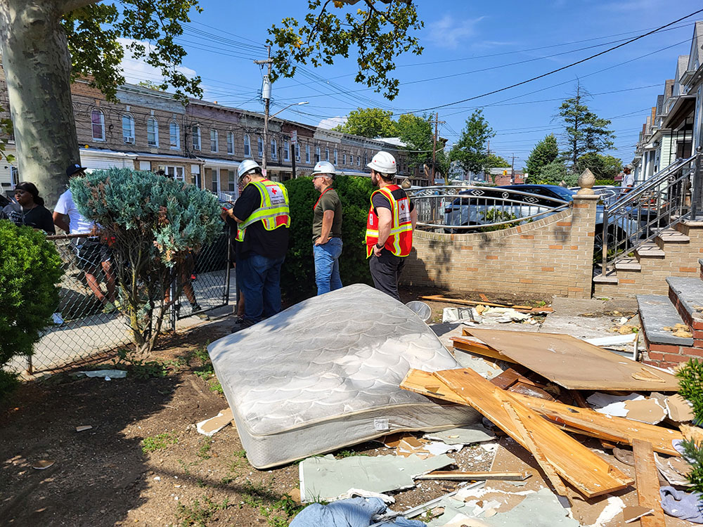 Three American Red Cross volunteers in vests and hard hats with a mattress and broken wood on the ground in a yard.