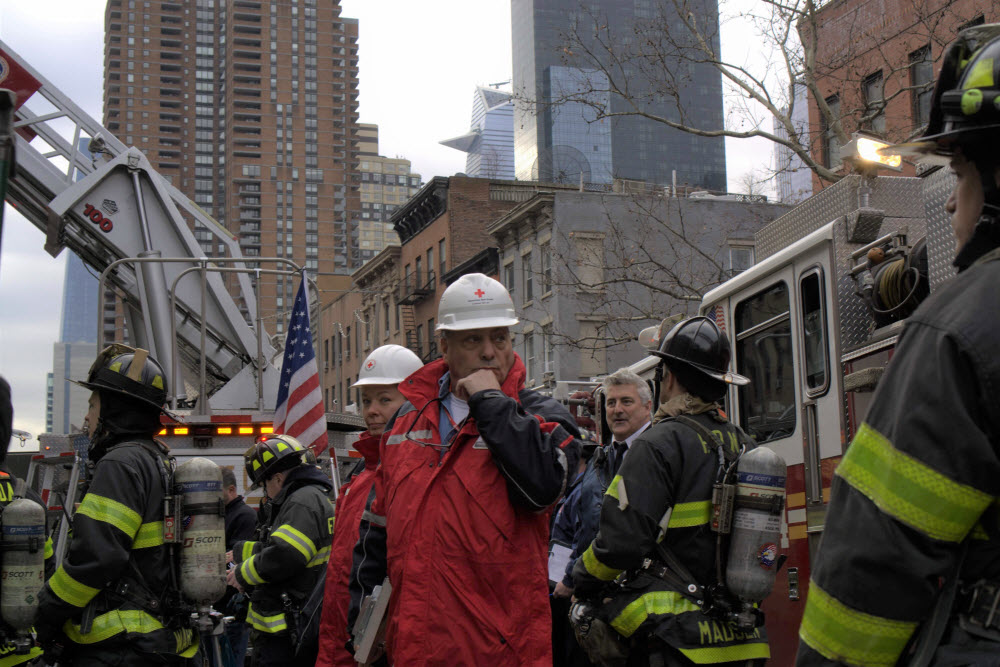 Volunteers in Red Cross jackets stand outside apartment building with New York City fire fighters.