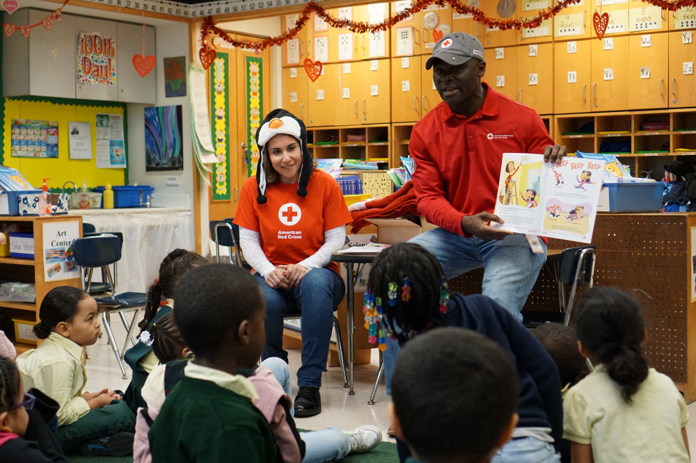 Red Cross volunteers wearing Red Cross T shirts sit in front of school children.