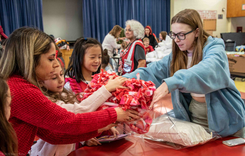 Red cross volunteers inside a school gymnasium packing blankets with elementary school children.