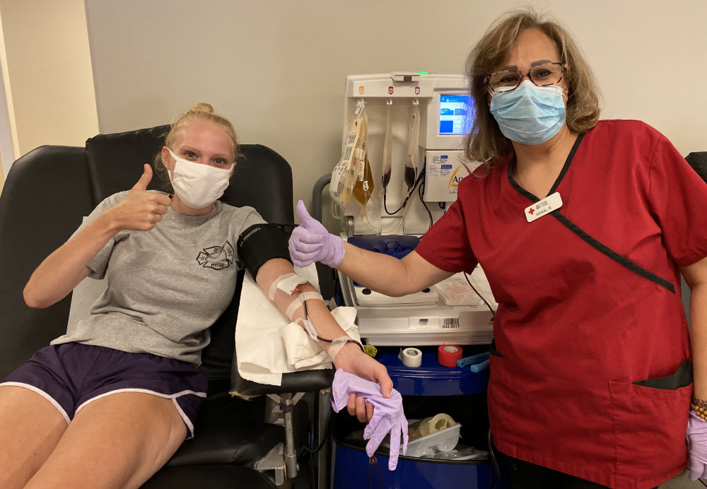 A young woman giving blood wearing a face mask and a Red Cross nurse.