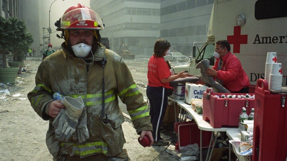 Firefighter in full gear walking through debris on streets and Red Cross volunteers with table full of supplies.