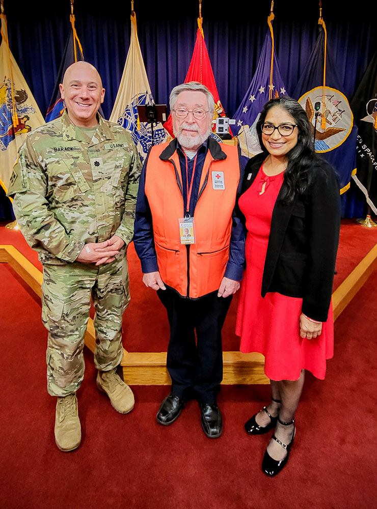 Red Cross volunteer John McGuire, Lt. Col. Matthew Barack, and and Lallita Maharaj in a room at Fort Hamilton with flags behind them.