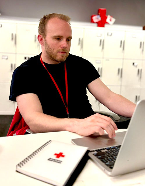 A man with short reddish hair wearing a black t-shirt sits at a table working on a silver laptop. A Red Cross-branded notebook lays next to him and a wall of lockers is behind him.