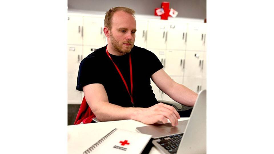 A man with short reddish hair wearing a black t-shirt sits at a table working on a silver laptop. A Red Cross-branded notebook lays next to him and a wall of lockers is behind him.