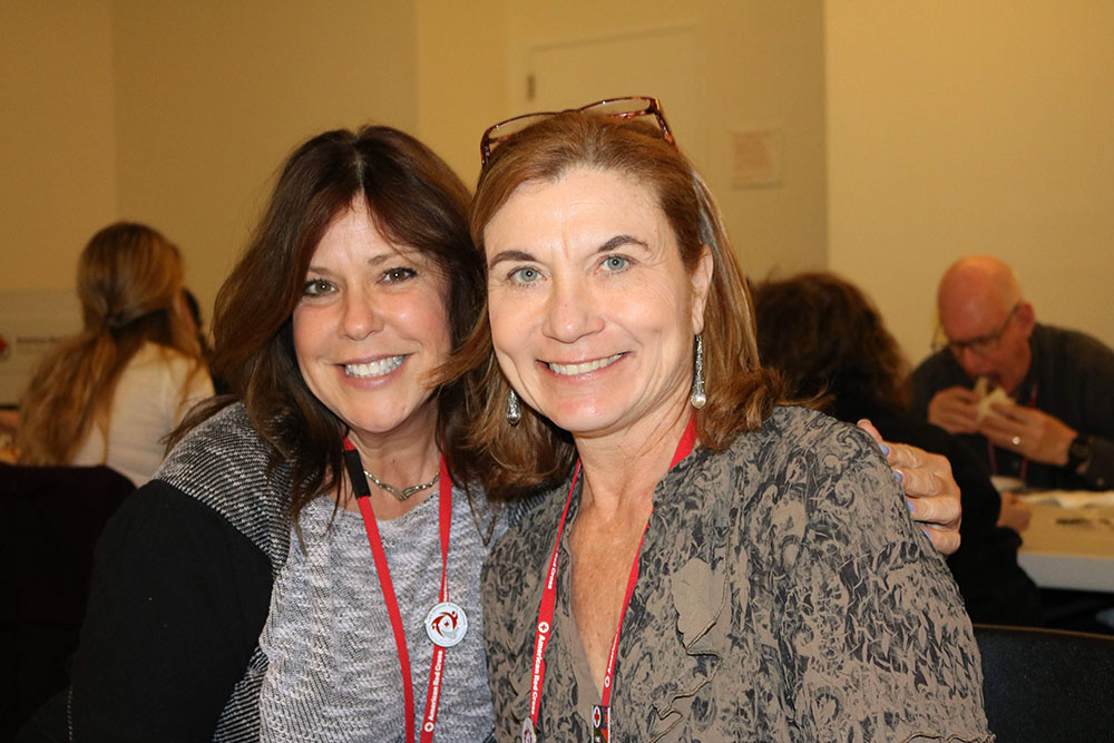 Two people pose next to each other wearing Red Cross lanyards around their necks. Behind them are other people in a blurry background.