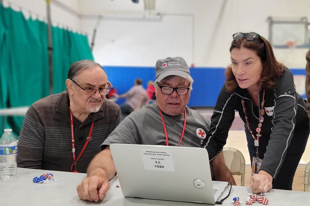Three people stand in a gymnasium space around a laptop computer. Each of the three people have Red Cross lanyards and other Red Cross logos on their clothes.