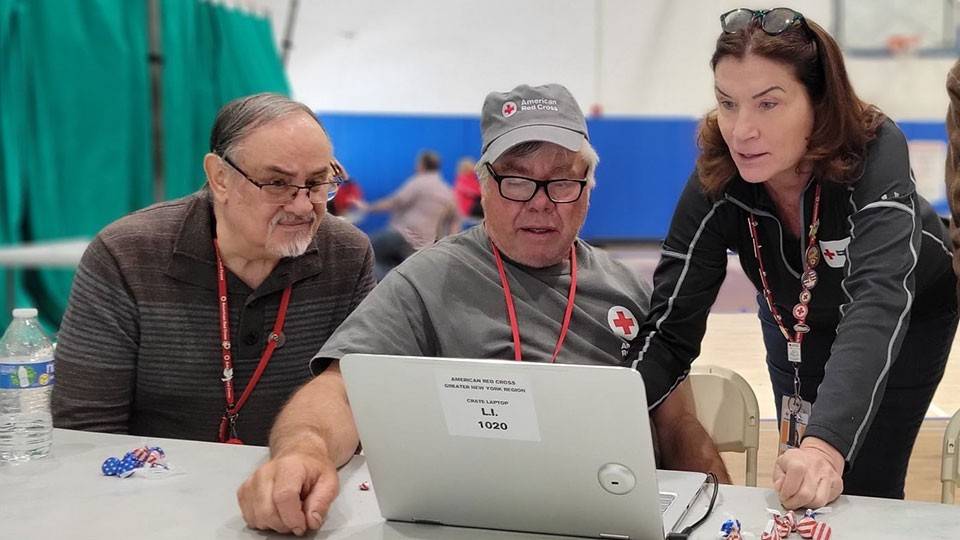 Three people stand in a gymnasium space around a laptop computer. Each of the three people have Red Cross lanyards and other Red Cross logos on their clothes.