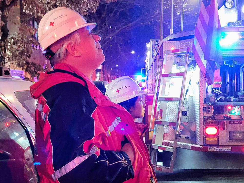 Red Cross volunteer Julio Medina wearing a Red Cross hard hat and vest, stands next to a fire engine with lights on that's next to an apartment building.