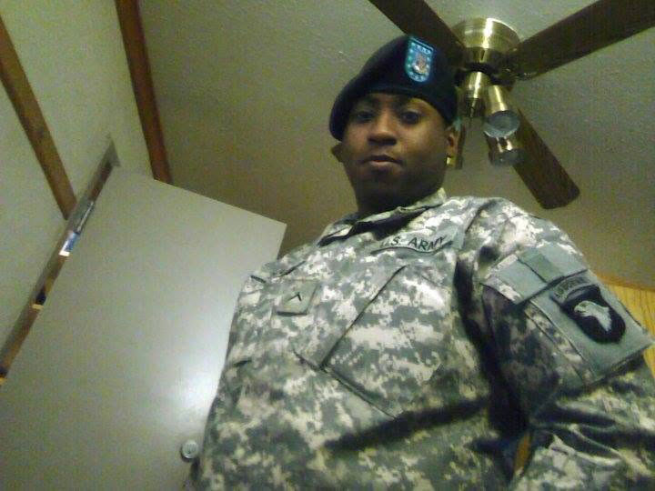 A man wearing Army fatigues and a beret with a blue crest on it. There is a patch on the shoulder. He is in a plain room with a ceiling fan above him and an open door behind him.