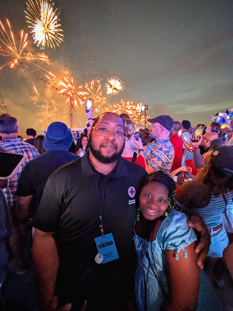 A man in a black polo with a Red Cross logo on the chest stands with his arm around his daughter wearing a blue dress and braids. There are fireworks in the sky above them and they are surrounded by people taking pictures of the fireworks.